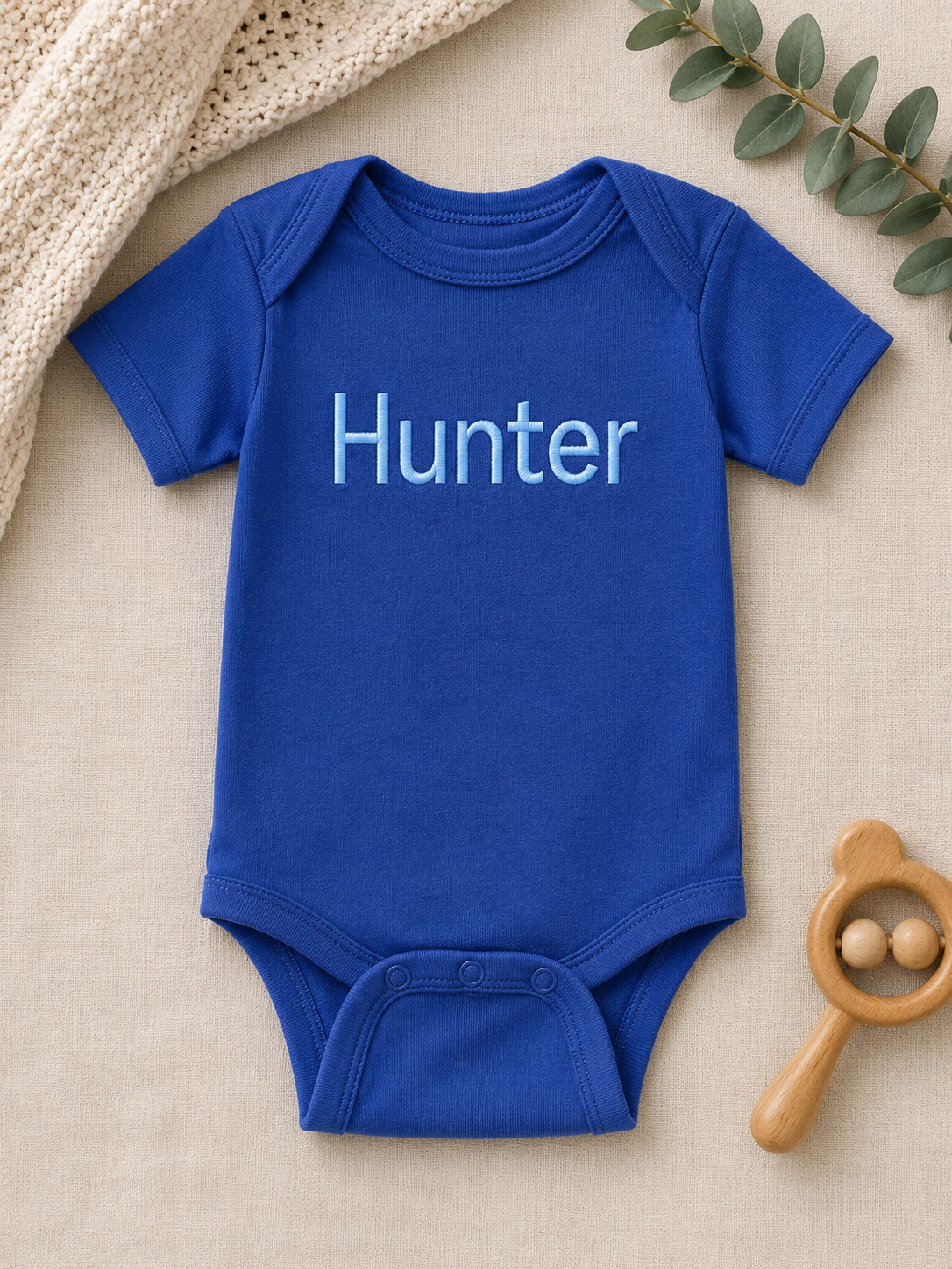 Blue baby onesie with 'Hunter' printed on it, placed on a beige surface with a wooden rattle and green leaves.