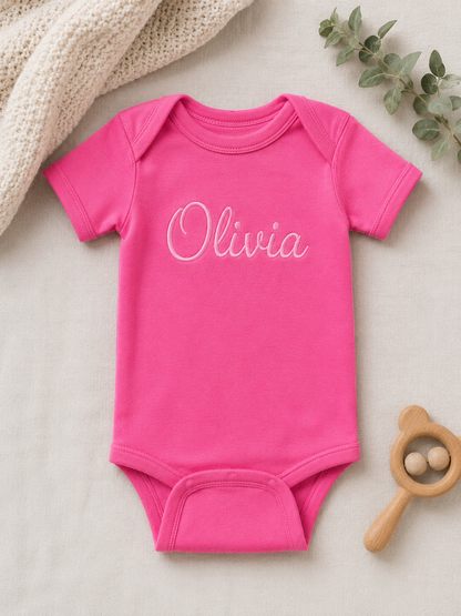 Pink baby onesie with baby's name embroidered on it, placed on a light surface with a wooden rattle and greenery.