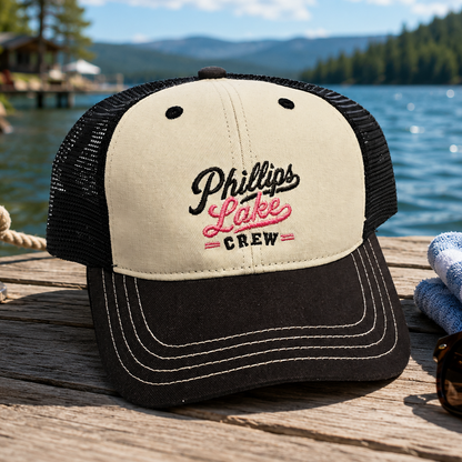 A trucker-style baseball cap with a cream front panel and black mesh back, featuring embroidered text reading 'Phillips Lake Crew' in black and pink. The cap is photographed on a wooden dock with a lake, trees, and mountains in the background.
