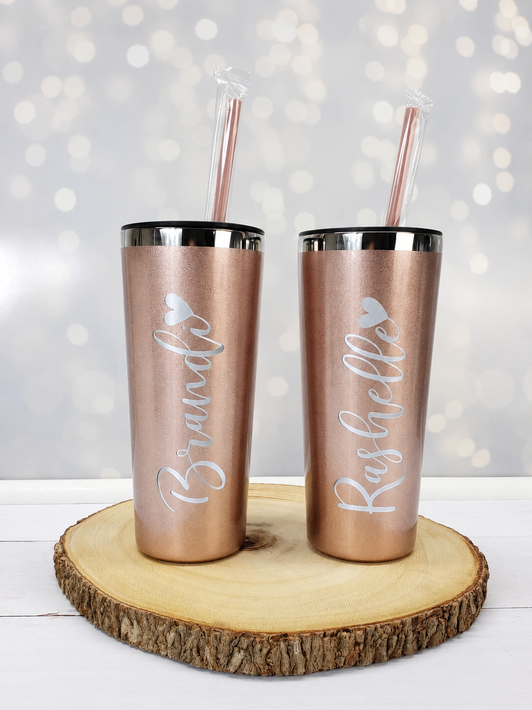 Two rose gold tumblers with names engraved on them, placed on a wooden stand with a blurred light background.