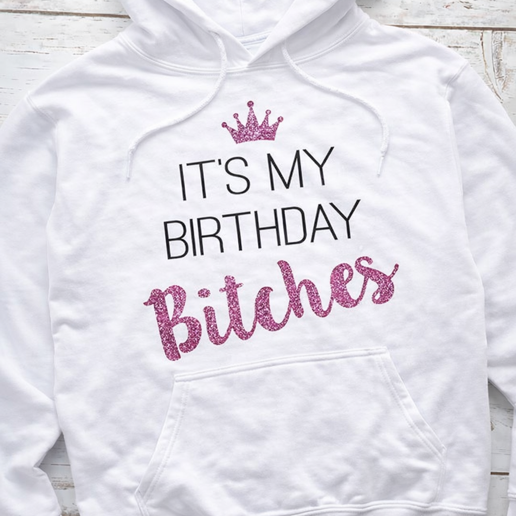 White hoodie with 'It's My Birthday Bitches' text in pink and black on a wooden surface