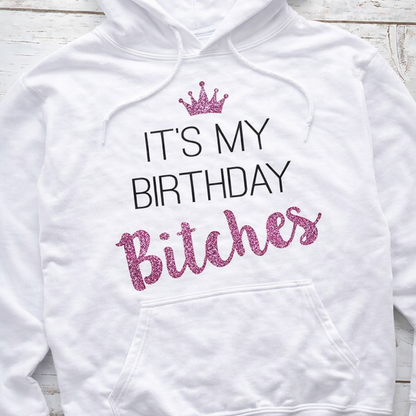 White hoodie with 'It's My Birthday Bitches' text in pink and black on a wooden surface