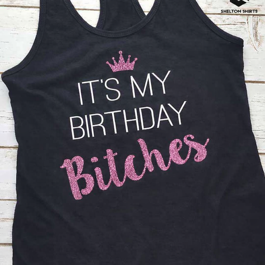 Black tank top with 'It's My Birthday Bitches' text in pink glitter on a wooden surface.