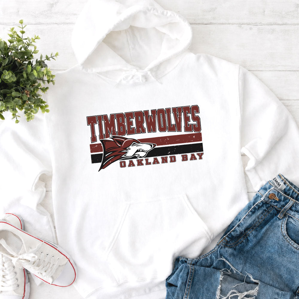 A white pullover hoodie laid flat on a light wood surface, featuring a distressed red, black, and white 'TIMBERWOLVES OAKLAND BAY' graphic on the chest. The hoodie is styled with blue jeans, white sneakers, and a small green plant.